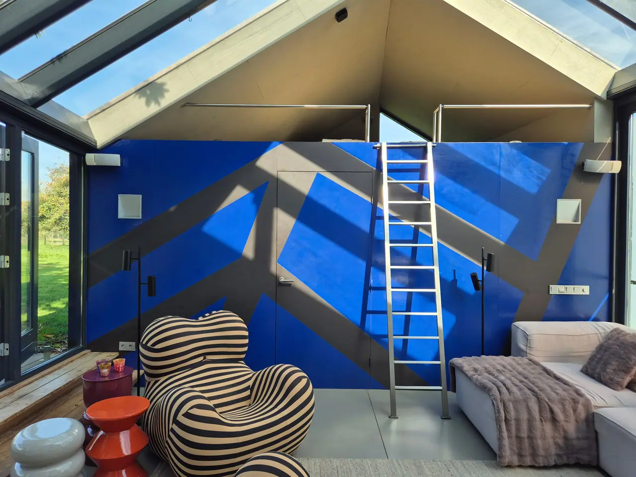 Woonkamer onder het glazen dak met kobaltblauwe wand, ladder naar de vide en gestreepte fauteuil